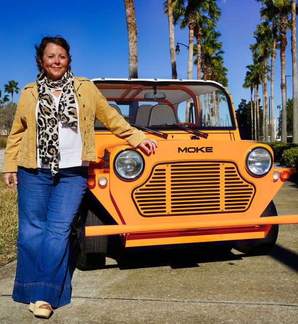 Suzanne standing next to an orange Moke car with palm trees and a clear blue sky in the background.