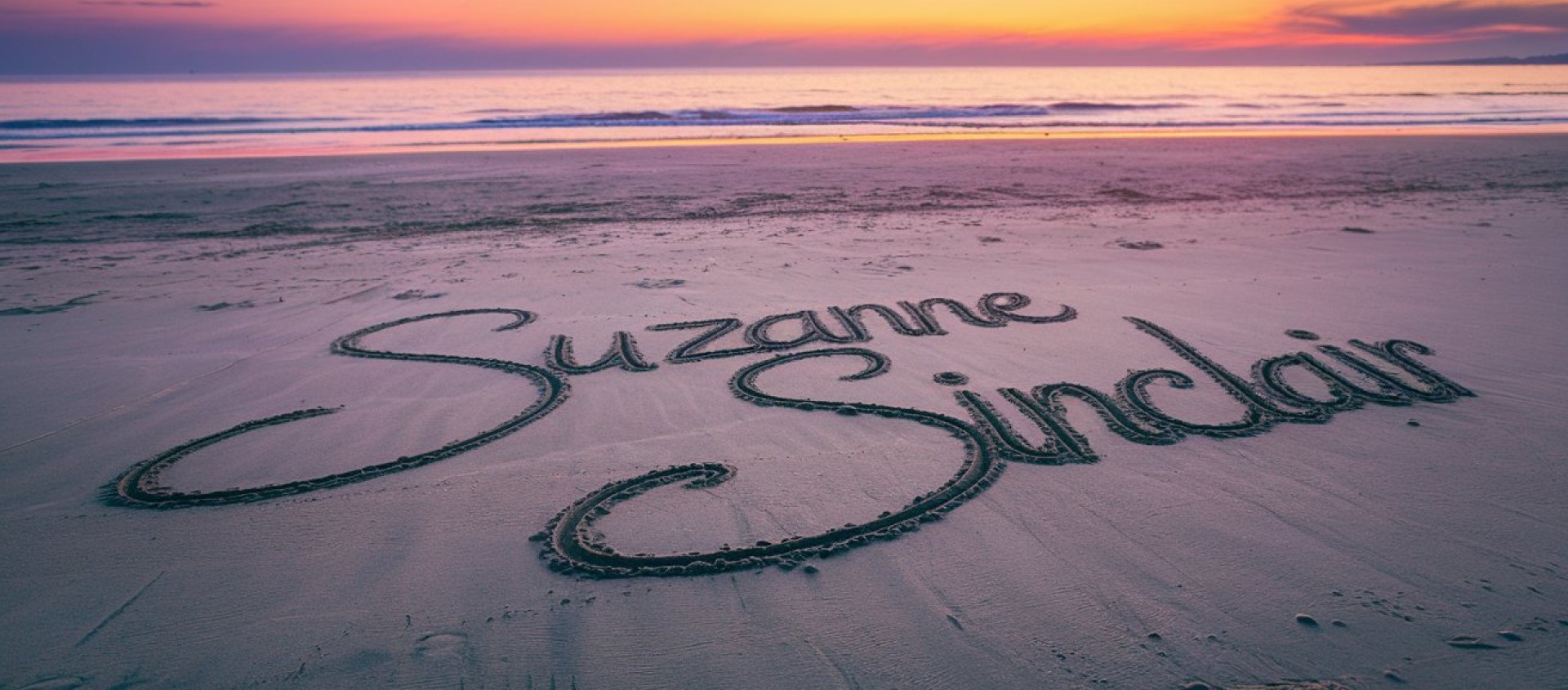 Name "Suzanne Sinclair" written in the sand on a beach at sunset